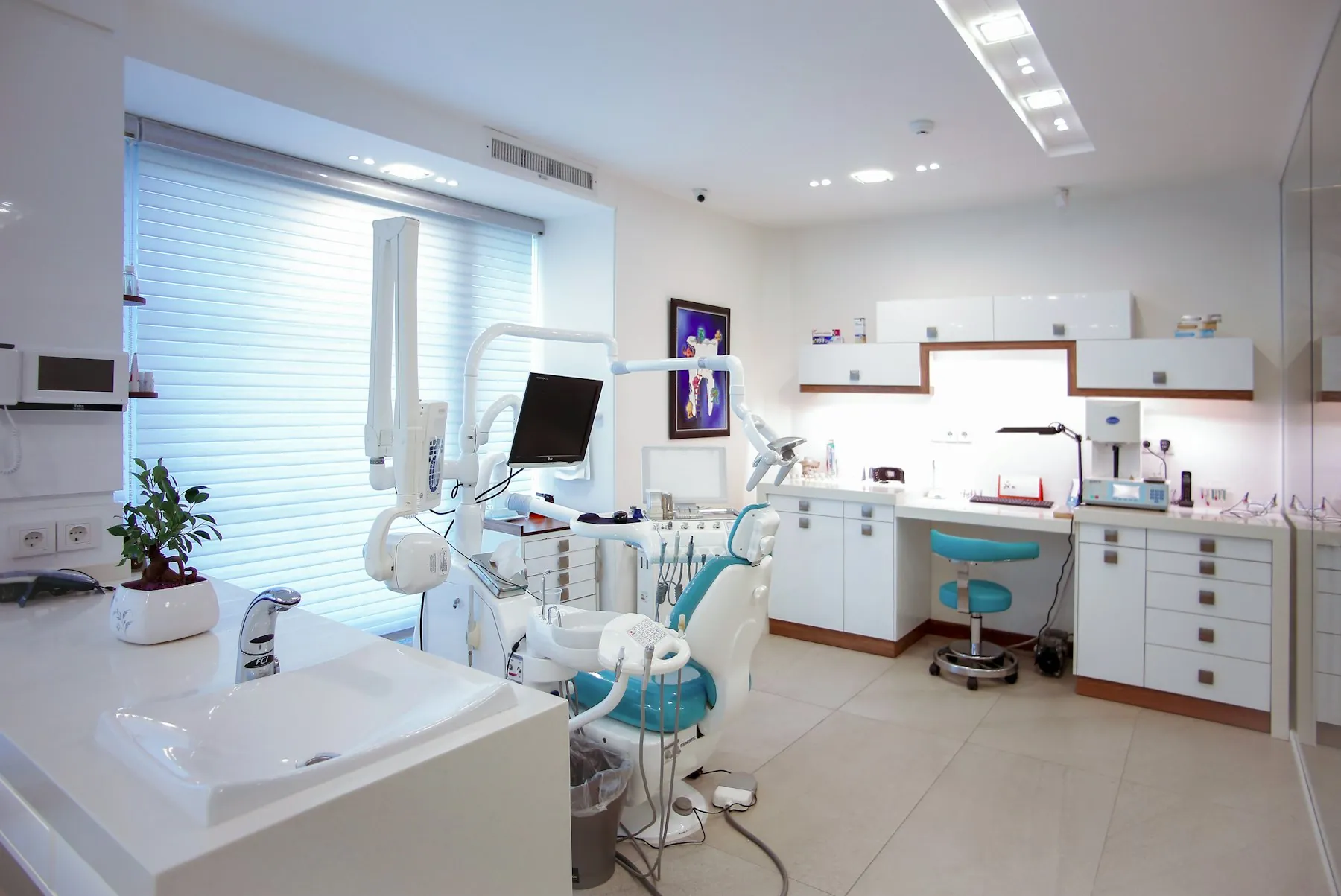 A bright, sea-glass toned dental treatment room with a chair, large windows and white cabinetry